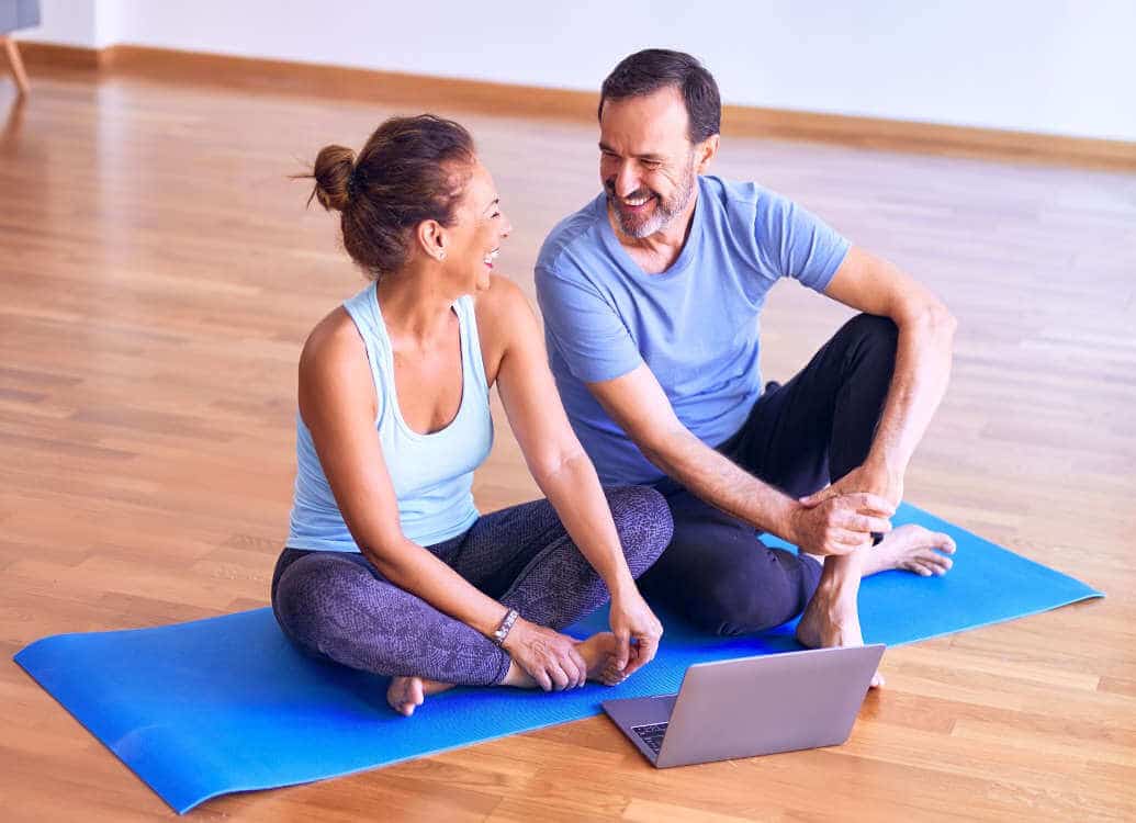 ein Mann und eine Frau, die auf einer Yogamatte vor einem Laptop sitzen