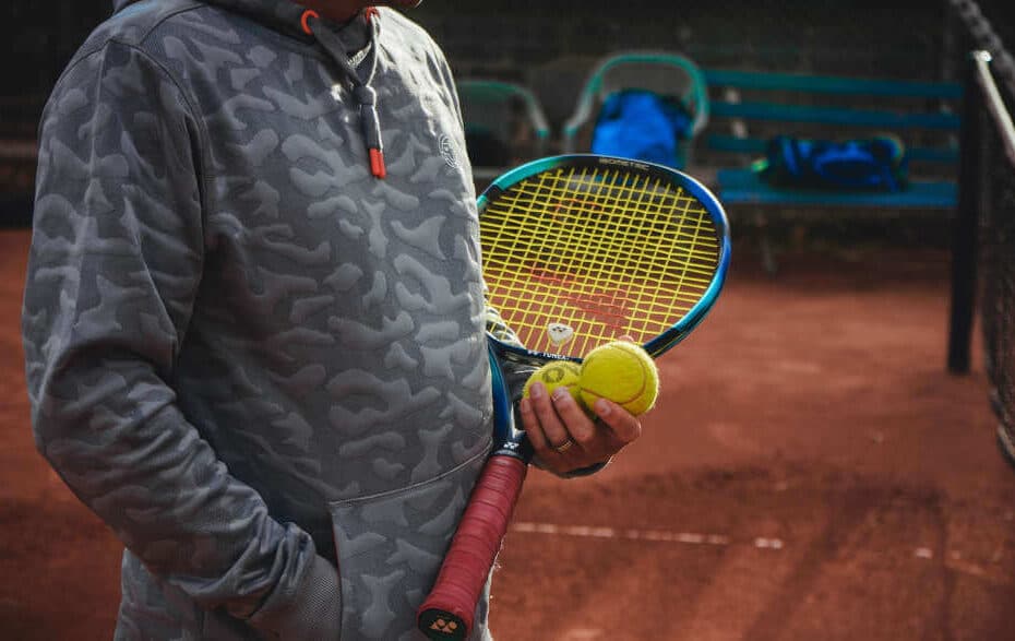 Tennistrainer steht auf einem Sandplatz mit drei Tennisbällen in der Hand und einem Tennisschläger