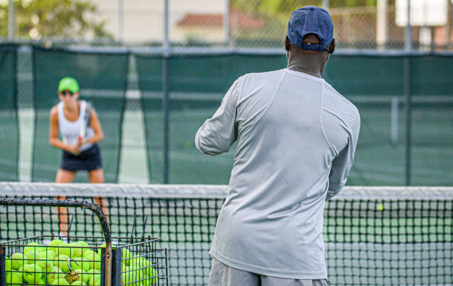 Tennistrainer mit einem Korb voller Tennisbälle und einem Tennisschüler, der auf einem Tennis-Hartplatz steht
