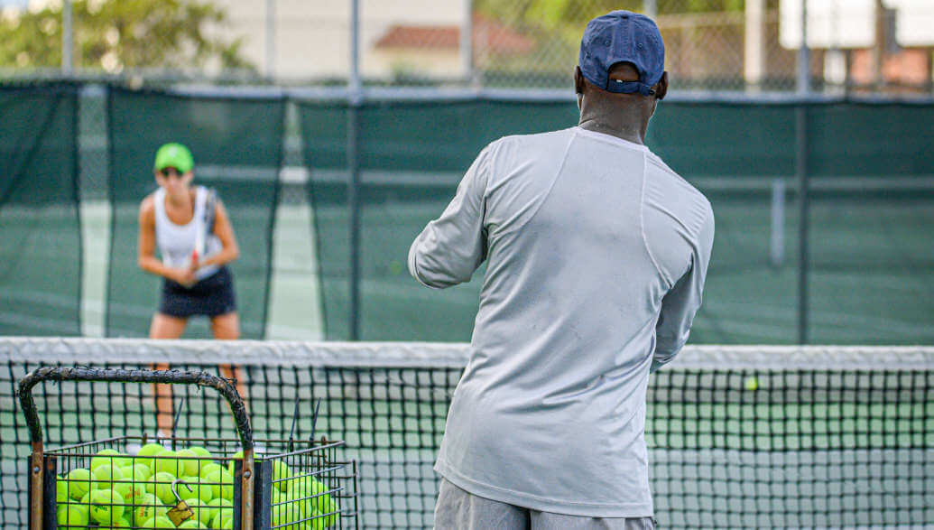 Tennistrainer mit einem Korb voller Tennisbälle und einem Tennisschüler, der auf einem Tennis-Hartplatz steht