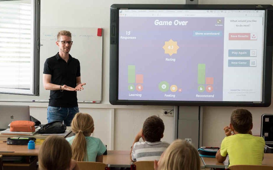 Ein Lehrer unterrichtet eine Grundschulklasse in einem Klassenzimmer, während er neben einem Bildschirm steht, auf dem ein Lernspiel angezeigt wird.