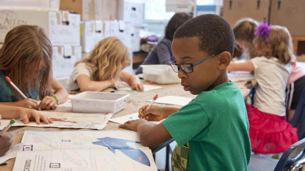 Six elementary school students draw on paper while sitting at their desks.