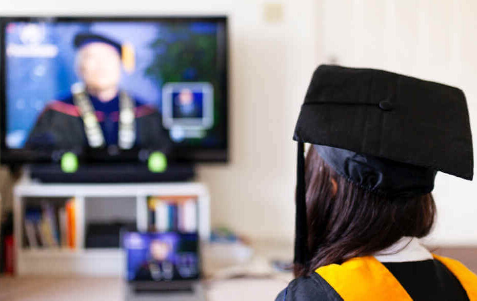 A woman in a graduation gown watching a graduation ceremony on TV.