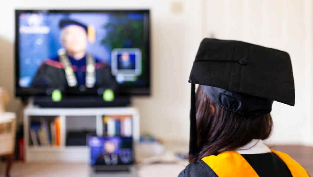 A woman in a graduation gown watching a graduation ceremony on TV.