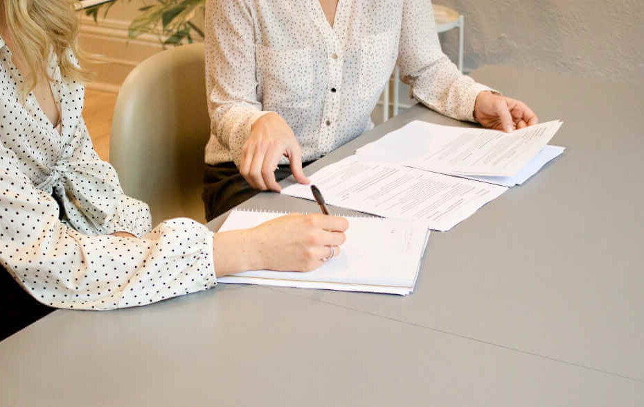 Two women are sitting at a table writing on a piece of paper.