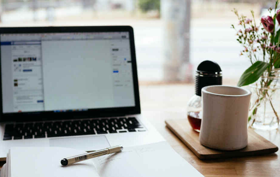 Ein Laptop, Blumen, ein Notizbuch und eine Tasse stehen auf einem Arbeitstisch.