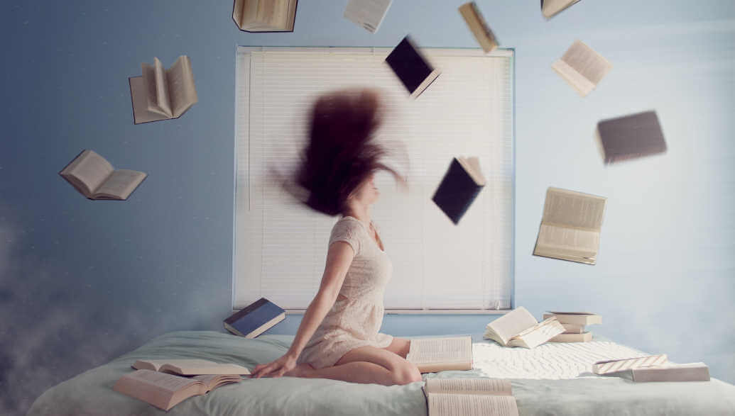 A woman is sitting on a bed throwing books in the air.