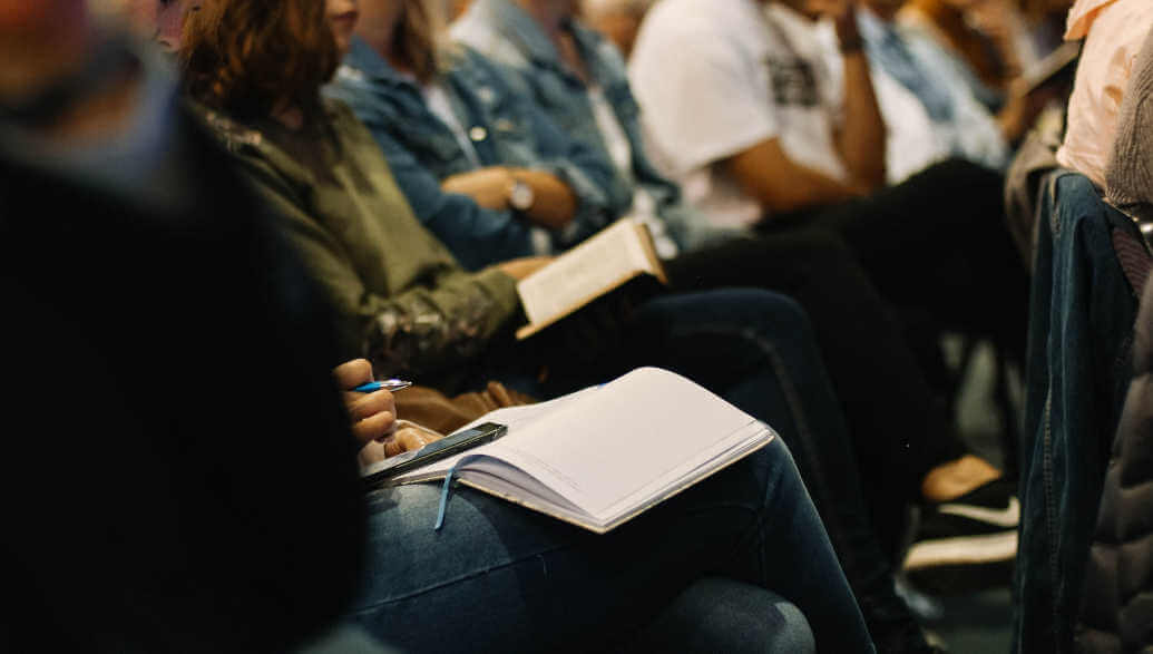 Many people sit on chairs with notebooks on their laps.