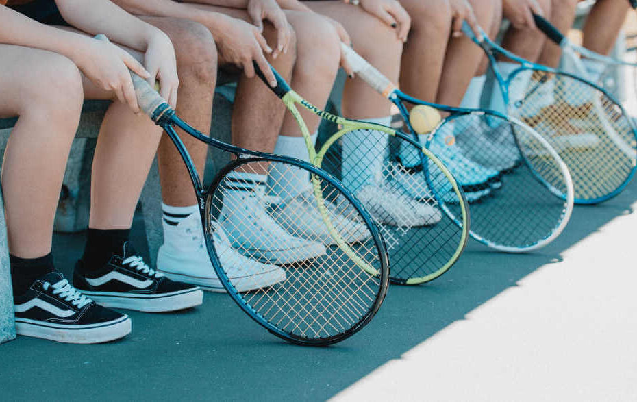 Six tennis players sit on a bench and hold their rackets in their hands.