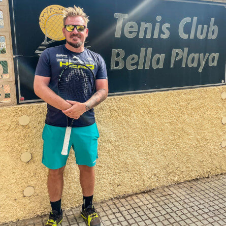 Tennis Coach Tim Haneklaus stands in front of a Tennis Club Bella Playa sign with his tennis racket in his hands.